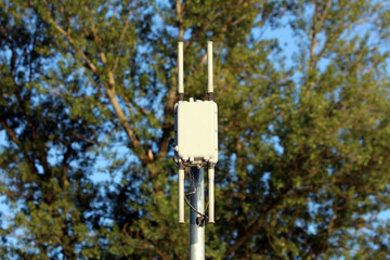 A small, box-like wireless access point with four short antennas is installed atop a metal pole, providing Wi-Fi or cellular signal distribution in a leafy suburban or park environment