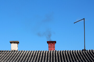 A red brick chimney sends black smoke into clear sky from a grey corrugated roof, indicating active...