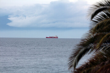 A large red oil tanker moves across calm open water beneath an overcast sky, with distant mountains...