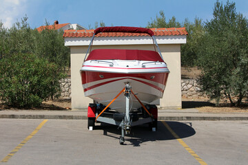 A white and red speedboat with a fitted red canopy is secured on a trailer in a marked parking...