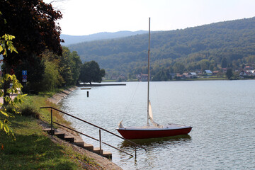 A small red sailboat rests by concrete lakeside steps, calm water reflecting village homes and...
