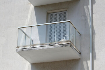 A compact residential balcony with glass railings, a white chair, and closed shutters receives...