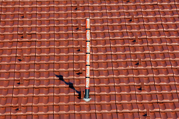 A vertical pole marked with numbers stands amid clean, red terracotta roof tiles, likely functioning as a snow depth or meteorological measurement tool under a clear sky