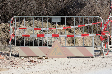 A metal barrier with red and white warning tape and a faded work sign blocks road access, warning...