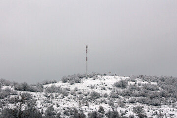 A slender telecom tower and a large cross stand atop a snow-covered hill, surrounded by...