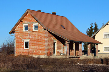 A partially constructed brick house with a sloped red tile roof stands in a grassy rural area, featuring exposed masonry and unpainted walls under a clear blue sky