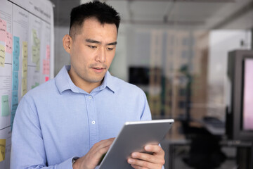 Focused Chinese businessman revieing ideas or plans displayed on tablet, accessing and learning digital information, looking up data, statistics, or research working on project stands in office. Tech