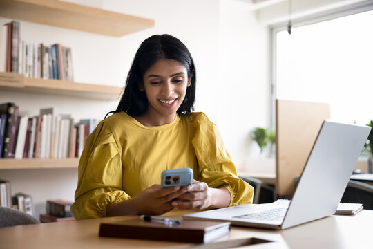 Smiling Indian woman using smartphone seated at workplace in office, lead positive interaction, enjoy personal communication, check email or messages, responding to professional correspondence via app - Powered by Adobe