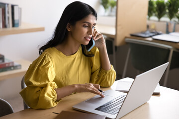 Serious Indian woman engaged in multitasking, speak on phone while working on laptop, discussing professional matter, managing project, coordinating tasks remotely, provide help to client by phonecall
