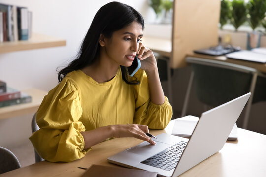 Serious Indian woman engaged in multitasking, speak on phone while working on laptop, discussing professional matter, managing project, coordinating tasks remotely, provide help to client by phonecall - Powered by Adobe