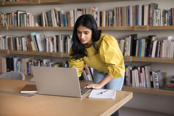 Focused Indian woman leaning at workplace desk using laptop, checking work updates or replying to...