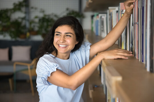 Cheerful young Indian woman engaged in selecting books for reading, studying, essay or exam preparation, standing nearby bookshelves of library, bookstore. Learning, passion for reading, new knowledge