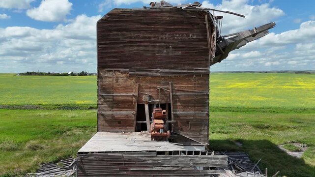 Large, dilapidated building sits in a field. The building is in a state of disrepair, with broken windows and a crumbling roof. The surrounding area is mostly grassy