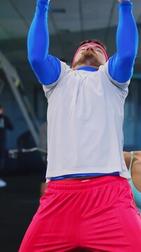 Funny cauxcasian baarded lazy man doing pull-up exercise with help of muscular athletivc fitness woman personal coach in the gym. Humor scene. Weak workout. Beginner concept.