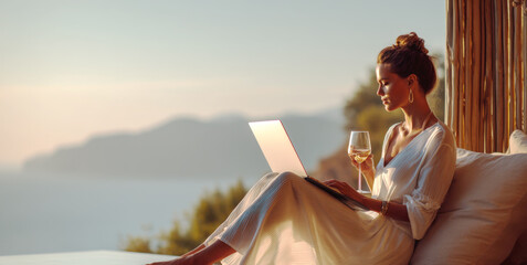 A stylish woman in a flowing white dress works on her laptop while enjoying a glass of wine by an infinity pool overlooking a serene ocean and mountains at sunset, embodying luxury remote work