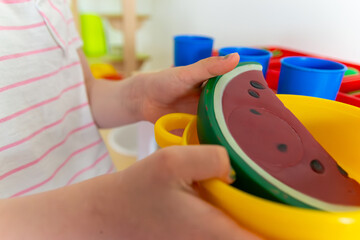 small child's hands holding toy watermelon slice in a bright yellow bowl, playing in a colorful preschool setting with plastic dishes, concept of early childhood education, concept of educational toys