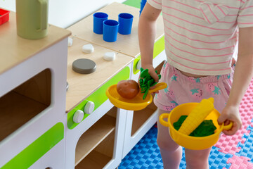 child playing with toy apple in a pan and green vegetables in a bowl near a wooden toy kitchen,...