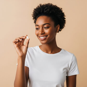 Happy healthy young adult African American woman holding vitamin in hand looking at pill standing isolated on beige background advertising beauty supplements nutrition treatment concept.