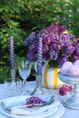 Beautiful lilac flowers, zephyrs, plates, cutlery, glasses and candles on table in garden, closeup