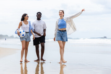 Three people enjoy summer outdoor activities on beach with people walking barefoot and holding ball...