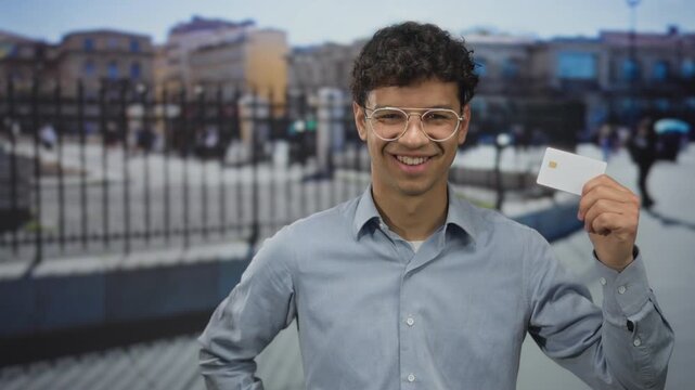 Young hispanic man holds blank card in right hand while smiling on busy city street near metal fence; confidence.