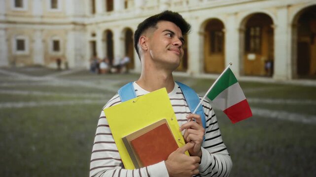 Italian student man smiles holding, books and italian flag in historic university courtyard.