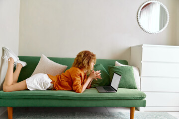 Young woman with curly hair using a laptop on a green sofa, enjoying her time at home, wearing...