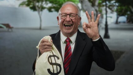 Senior man in a business suit outdoors holding a money bag with a dollar sign, smiling and showing the ok gesture on a street, implying themes of success or corruption.