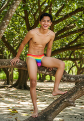 Happy young southeast asia man in colorful swim briefs sitting on a tropical tree at the beach. Ideal image for summer holidays, beach travel, relaxation, freedom, and vibrant outdoor lifestyle themes