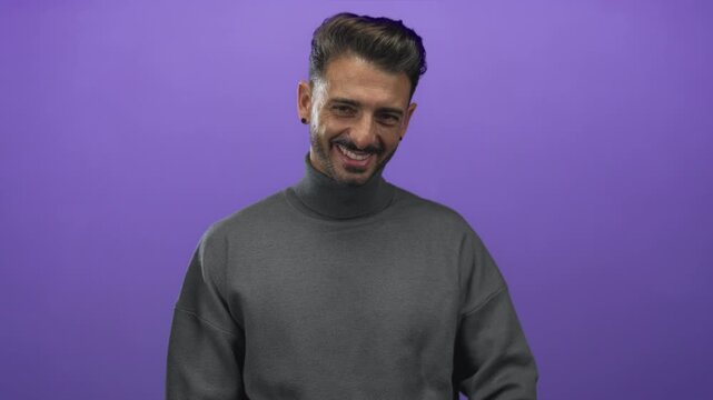 Young hispanic man wearing grey turtleneck and beard smiles and points finger to head in studio; confidence.