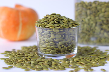 Glass jar filled with green pumpkin seeds or pepitas on a white surface, surrounded by scattered...