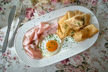 A Delicious Breakfast Plate Featuring Freshly Cooked Eggs, Tender Ham, and Fluffy Crepes Decorated with Chopped Herbs on an Elegant Floral Tablecloth