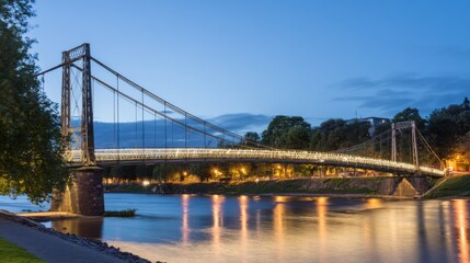 A suspension bridge illuminated at dusk with visible cable system, steel towers, warm lights reflecting on the river,
