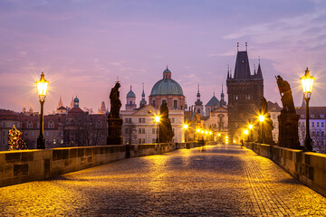 Obraz premium Charles Bridge at twilight in Prague, Czech Republic, beautifully illuminated with golden streetlights and surrounded by historic baroque architecture under a soft purple sky.
