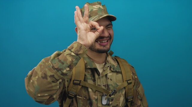 Man standing in camouflage uniform wearing backpack straps and dog tags shows ok sign in studio; confidence.