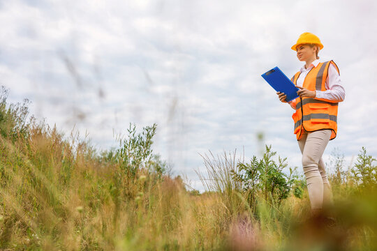 A female engineer wearing a safety vest and yellow helmet communicates via mobile phone while holding a clipboard, standing outdoors in a grassy area