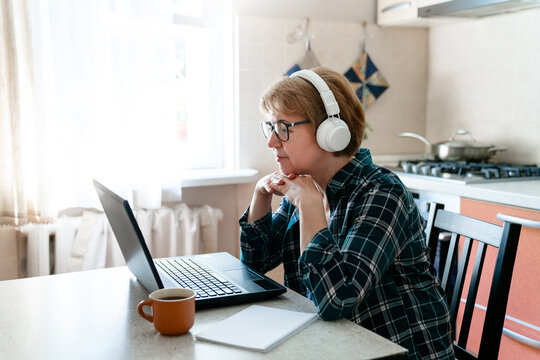Middle-aged woman in headphones working from home kitchen with laptop. Distance senior inclusive education, video call, therapy online. E-learning, digital communication. Technology lifestyle