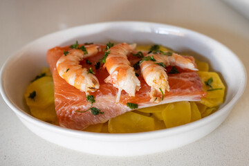 Fresh salmon fillet seasoned with parsley and topped with shrimp, resting on sliced potatoes in a white ceramic dish, creating a balanced, nutritious meal rich in omega-3s and proteins
