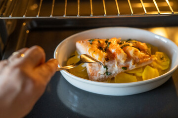 Cook uses a fork to check the doneness of a salmon fillet with potatoes, seasoned with herbs and spices, baking in a white ceramic dish in a modern oven