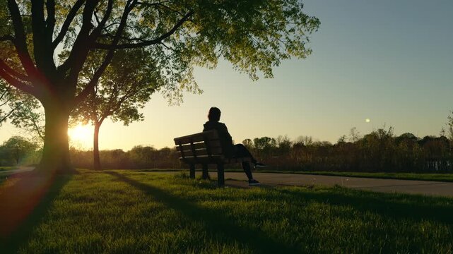 A silhouette of a woman sits on a bench and rests, sunset time. 
