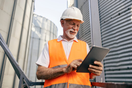 Portrait of an old refinery worker standing outdoors surrounded by silos and using tablet.