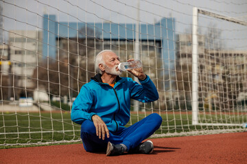 Tired thirsty senior sportsman sitting with crossed legs on stadium and drinking water.