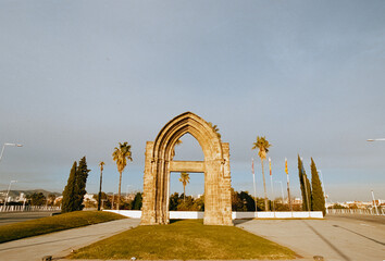 A color analog 35mm film photograph captures an ancient stone arch monument in a Barcelona roundabout, surrounded by palm trees under a bright sky, with classic film grain.