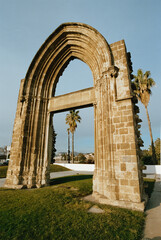 Fototapeta premium A 35mm analog photograph captures an ancient stone arch monument in Barcelona, showcasing its historic texture and details under a clear sky with palm trees.