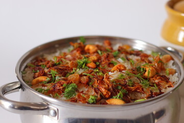 Indian chicken biryani served in a stainless steel pot, topped with fried onions, cashews, raisins, and fresh cilantro