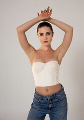A woman wearing a white corset and blue jeans poses with her arms raised against a plain...
