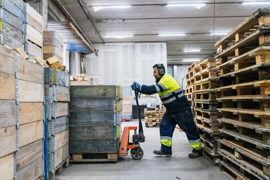 Warehouse worker transporting wooden pallets using manual pallet jack in a recycling factory, demonstrating efficient material handling and sustainable practices