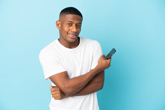 Young latin man isolated on blue background holding a mobile phone and with arms crossed