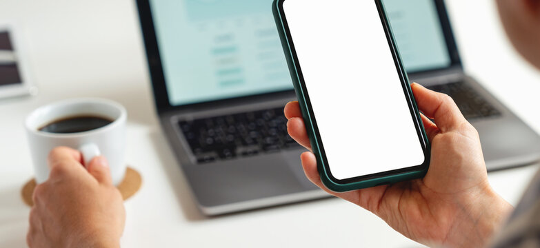 Mockup of smartphone with white blank screen in hand of businesswoman against background of laptop on table