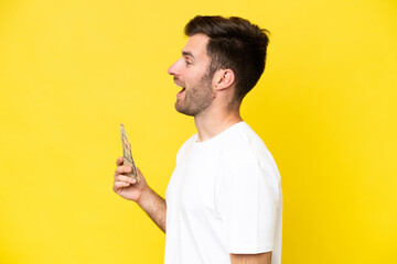 Young caucasian man taking a lot of money isolated on yellow background laughing in lateral position